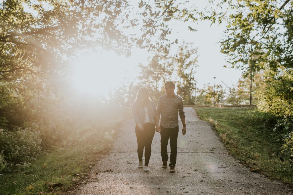 a man and a woman backlit by the sun walk down a path while holding hands during their Eagle Creek Engagement Photos