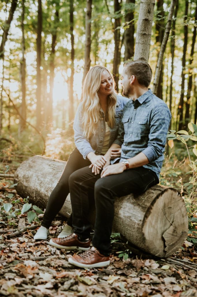 a blonde woman sits on a log with a bearded man while backlit by the sun streaming through the trees