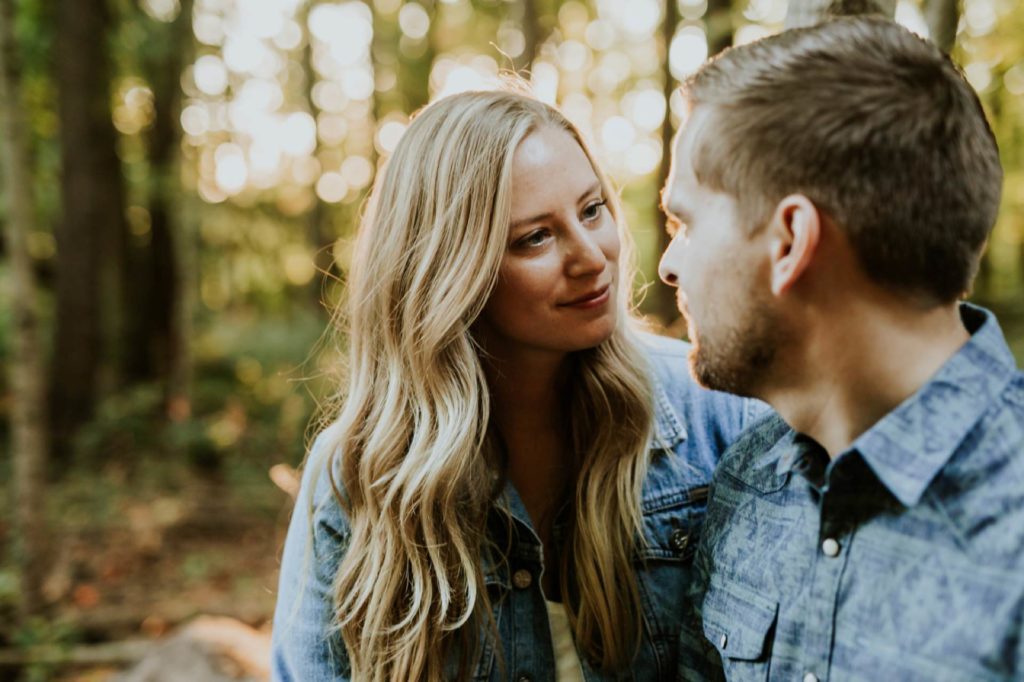 a blonde woman looks at a bearded man in the woods and smiles at sunset during their Eagle Creek Engagement Photos