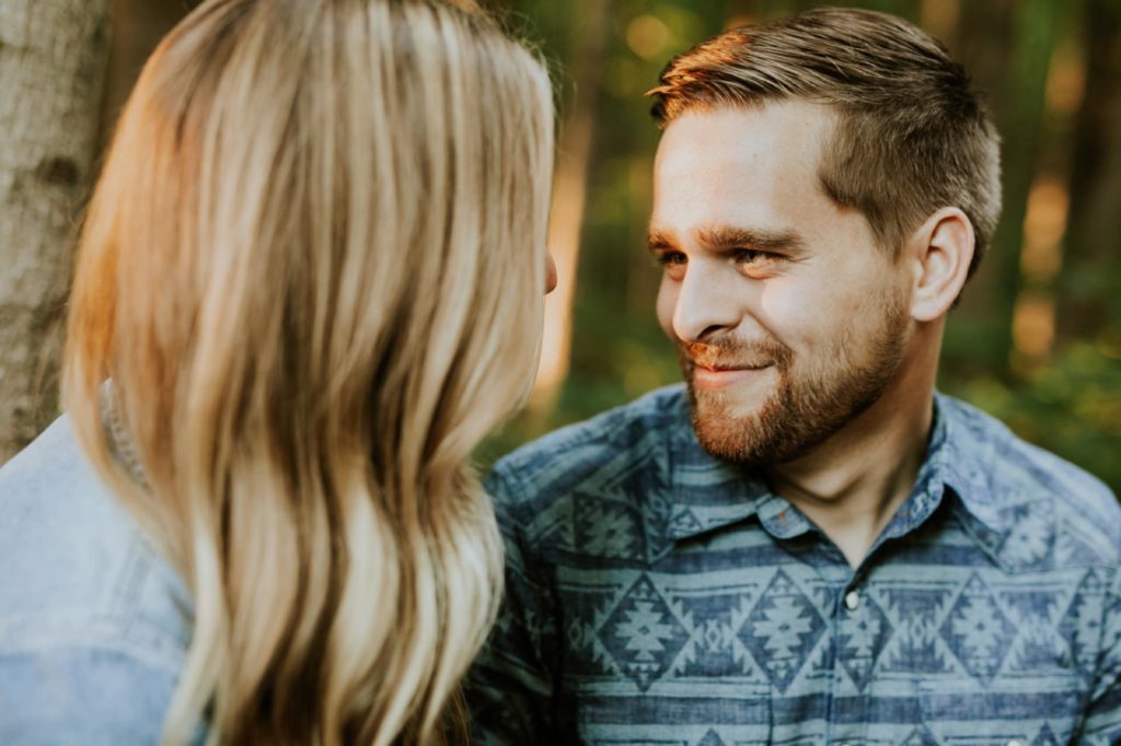 a bearded man looks at a blonde woman and smiles in the woods during their indy park engagement