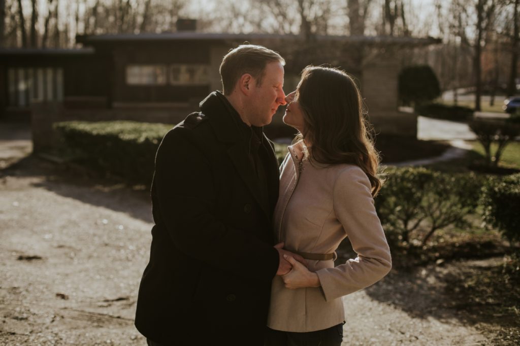 a man and a woman, both in jackets for chilly weather, kiss with the sun backlighting them at this Holcomb Gardens Winter Engagement