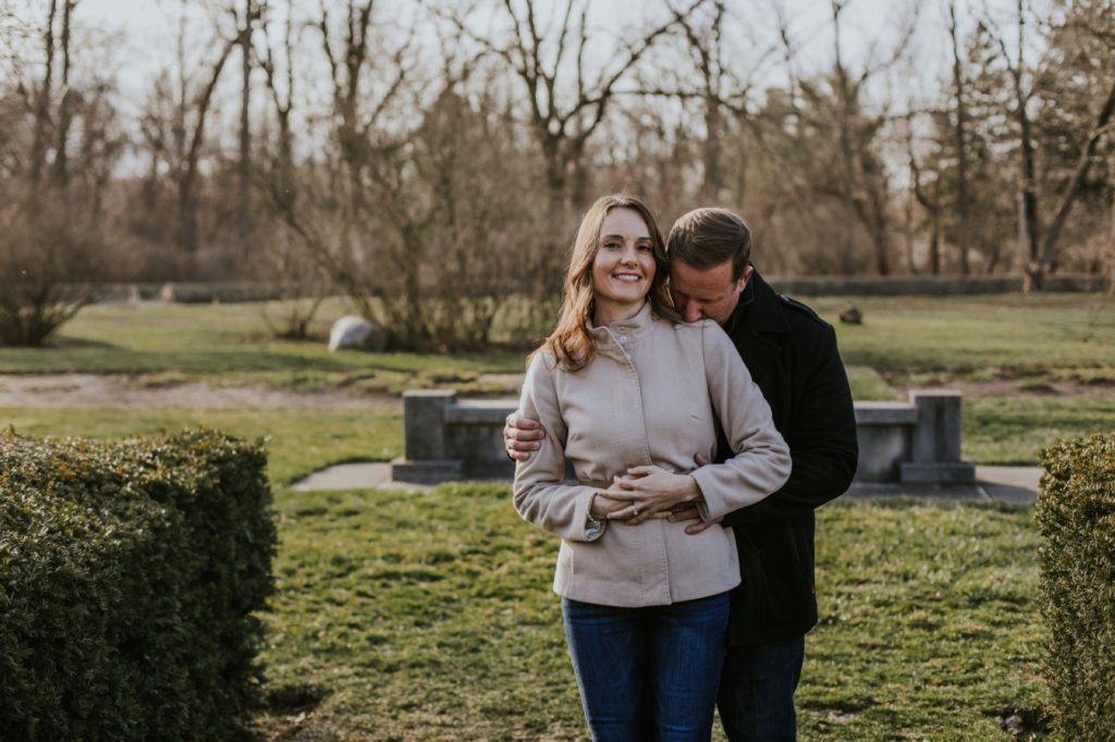 man in jacket kisses woman in jacket on shoulder in their holcomb gardens winter engagement shoot