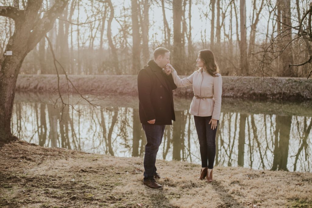 a man kisses a woman's hand in front of a canal during their holcomb gardens winter engagement shoot