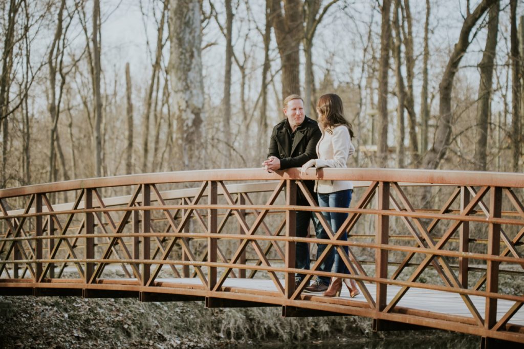 a man and woman stand on a bridge and chat during their holcomb gardens winter engagement shoot