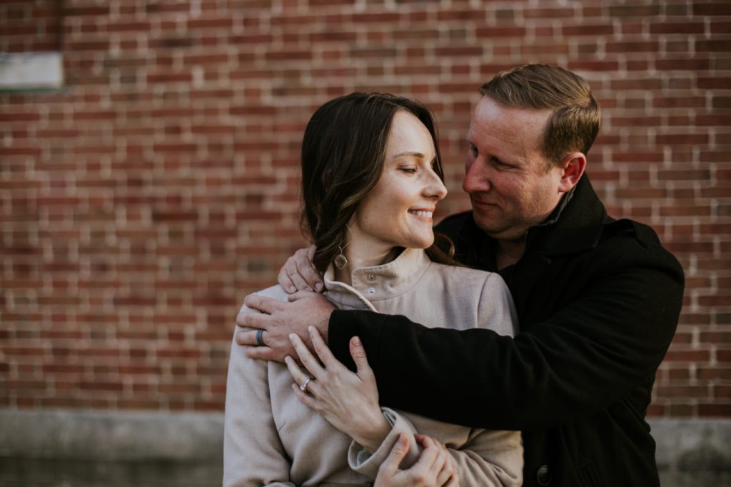 a man and a woman in jackets on a chilly day hug in front of a brick wall during their butler university portrait session