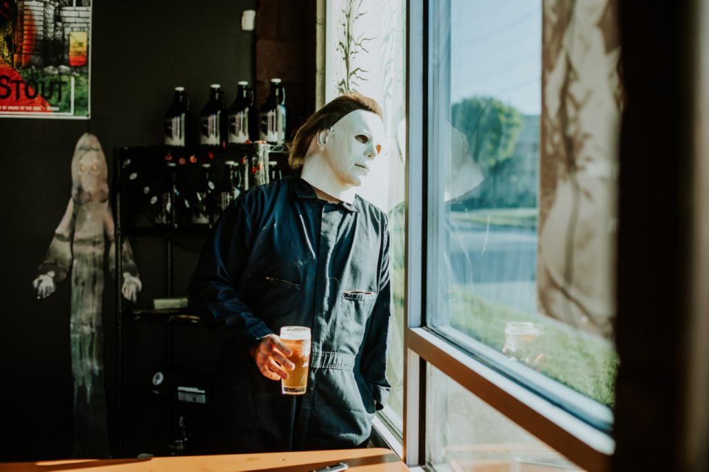 man with white face mask holds a beer and looks out the window