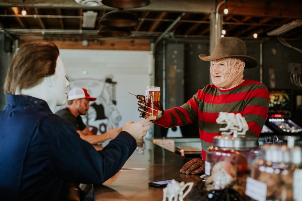 man with scarred face and claws and stripey shirt drinks beer with man with white face mask on in this Indianapolis brewery photography