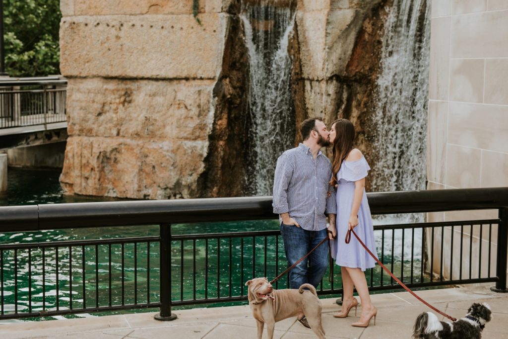 man and woman kiss in front of water fall in white river state park engagement picture while holding two dogs on leashes