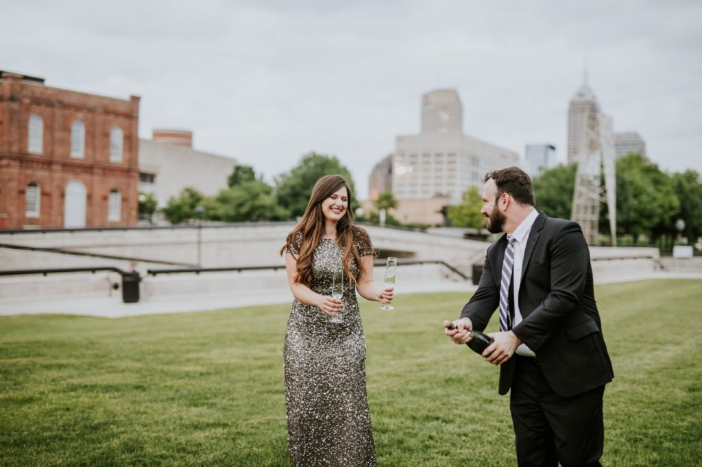 bearded man in suit pops a bottle of champagne in white river state park engagement picture