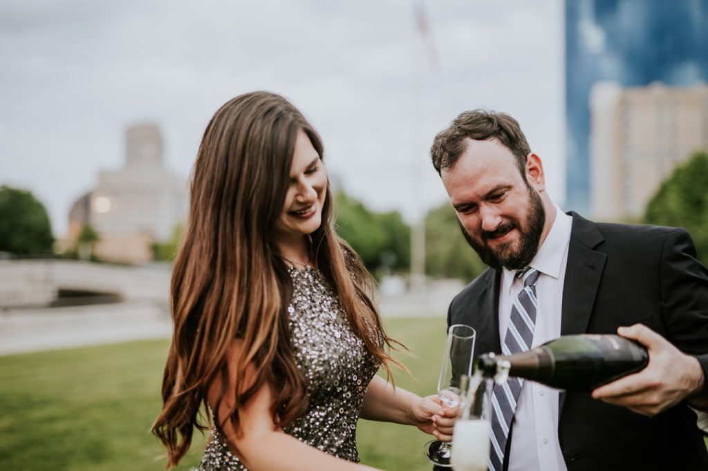 man in suit pours champagne into flutes with woman in dress in white river state park engagement picture