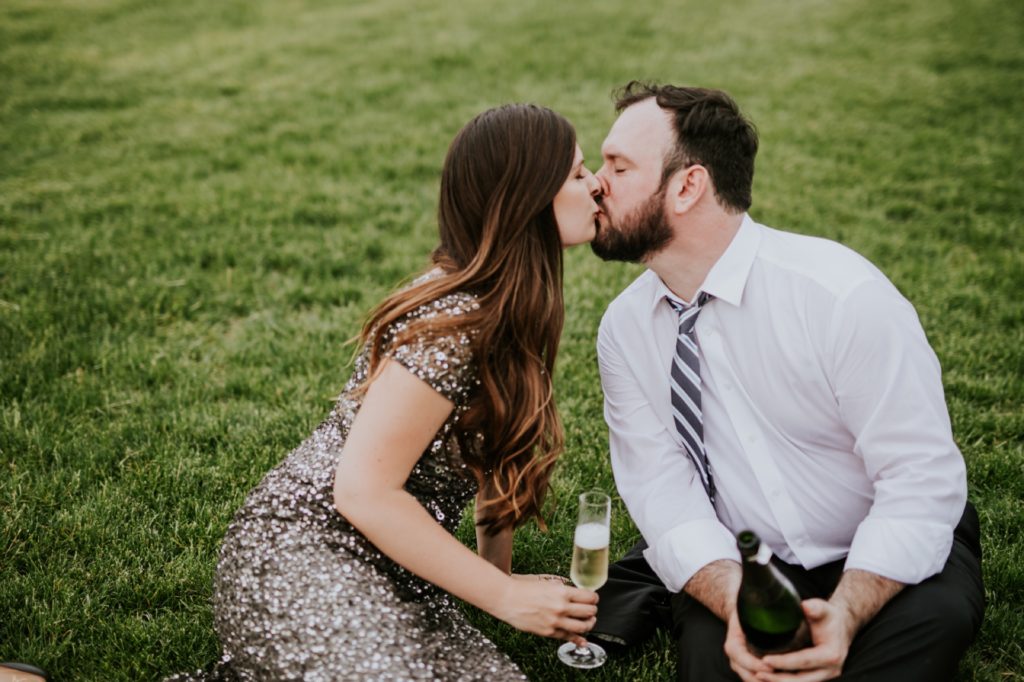 man and woman in formal wear kiss while sitting in grass in white river state park engagement picture