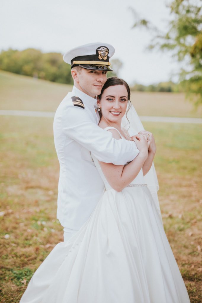 man in naval full dress whites hugs bride from behind in jackson morrow park