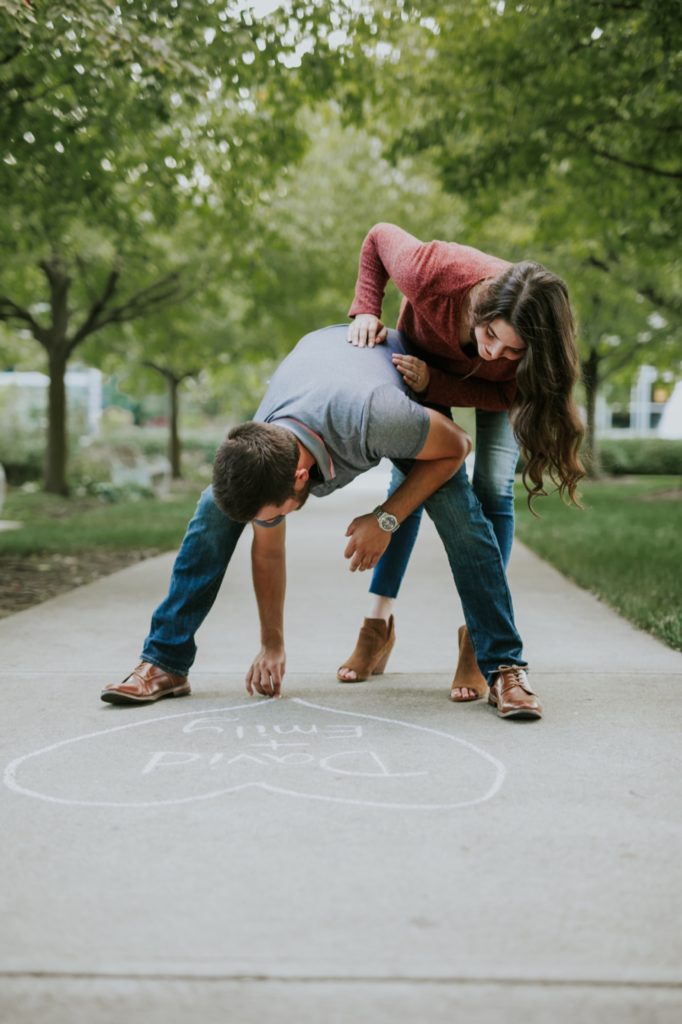 woman in red shirt looks over man in gray polo's shoulder as he draws with chalk on the ground during their newfields portrait session