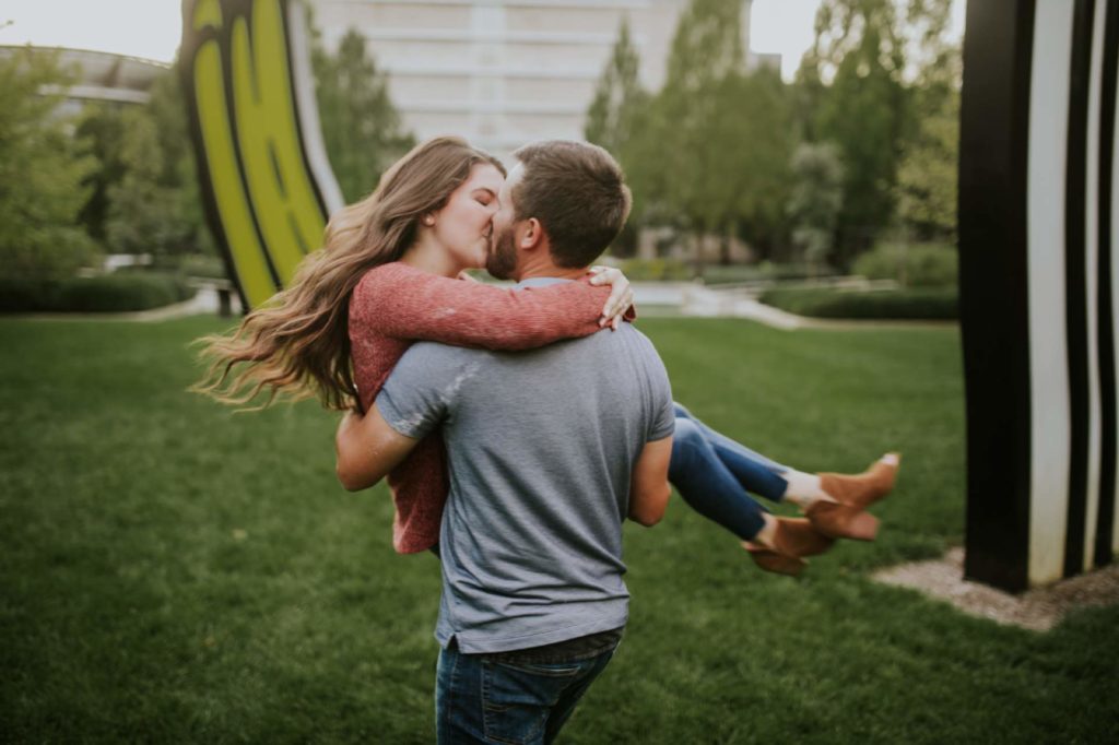 man in gray polo kisses woman in red shirt as he holds her in his arms and spins around during their IMA engagement session