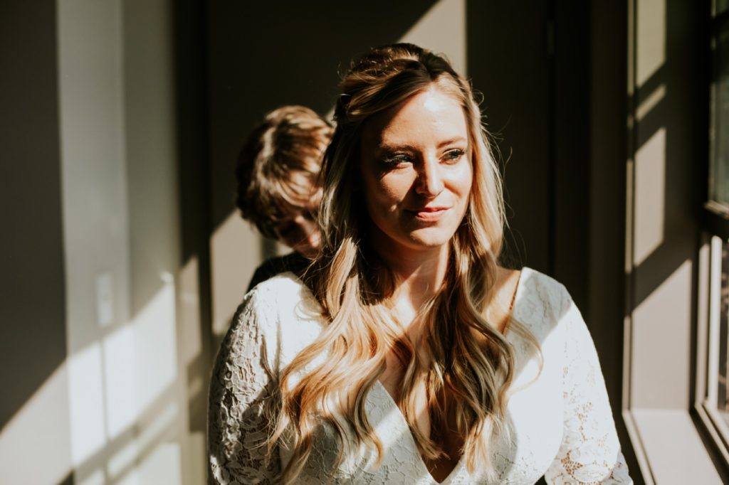 bride stands in window light while putting on her dress in blumlux building in indianapolis
