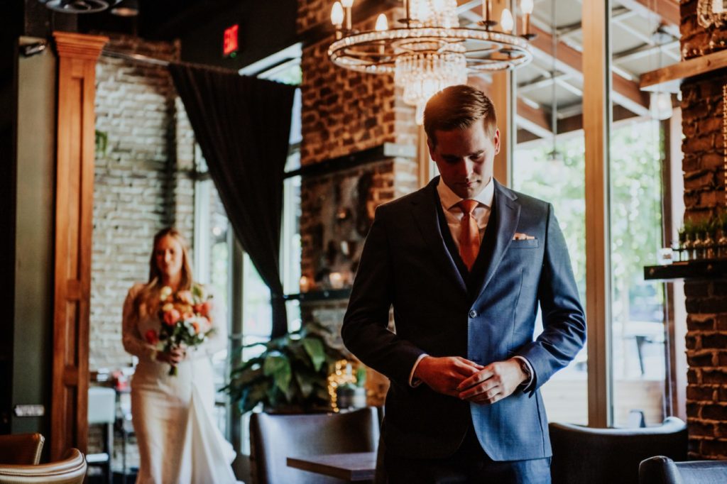 groom in blue suit stands under chandelier at the ball and biscuit while his bride enters the building behind him