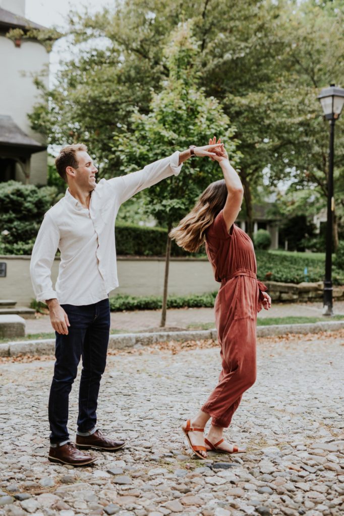 man in white shirt spins woman in orange jumpsuit on historic lockerbie street, one of the Best Indianapolis Engagement Photo Locations