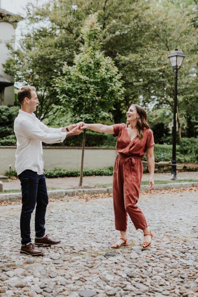 man in black pants and white shirt dances with woman in orange jumpsuit on historic lockerbie street, one of the Best Indianapolis Engagement Photo Locations
