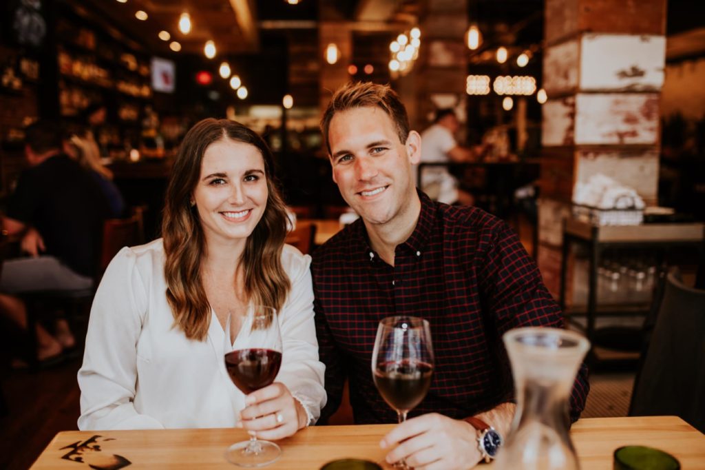 woman in white shirt and man in black drink wine for Louies Wine Dive engagement portraits