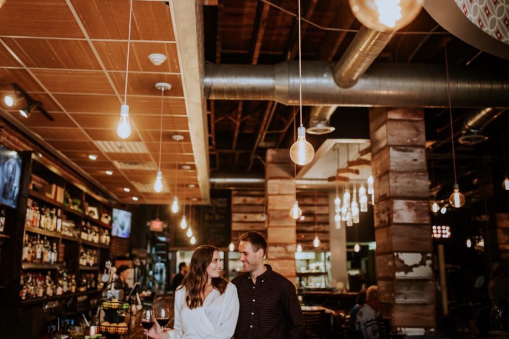 woman in white and man in black stand under hanging tungsten bulbs in Louies Wine Dive engagement portraits
