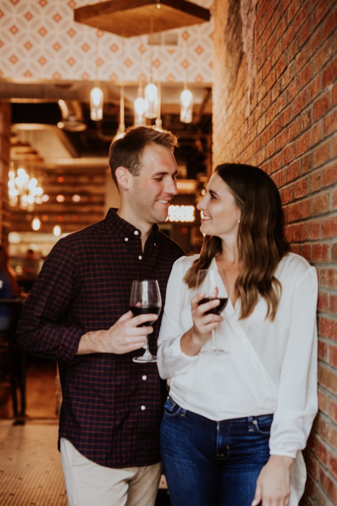 woman in white shirt leans against brick wall during Louies Wine Dive engagement portraits with her fiance