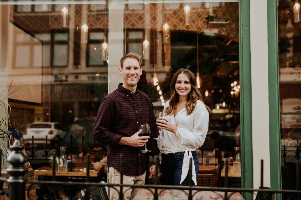 man and woman hold wine and drink wine outside Louies Wine Dive for their engagement portraits