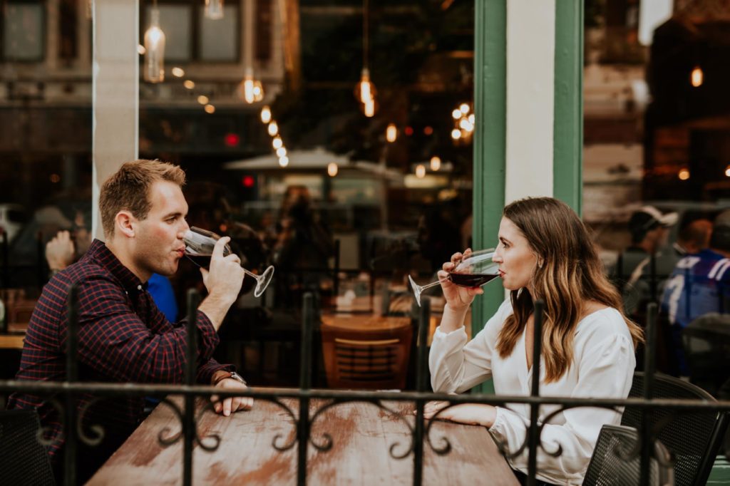 man and woman sit behind spiky black metal fence drinking wine for their Louies Wine Dive engagement portraits