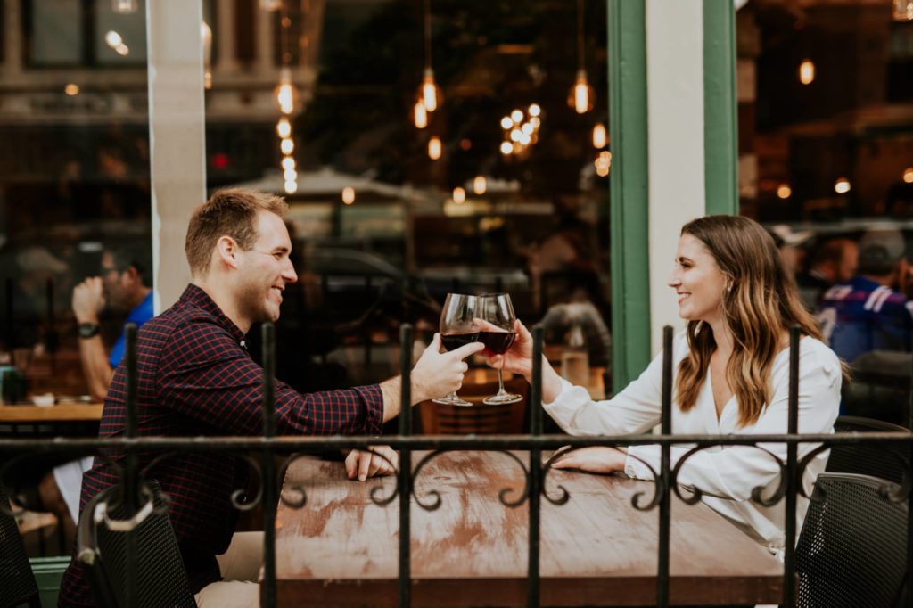 man in black and woman in white clink wine glasses outside during their Louies Wine Dive engagement portraits