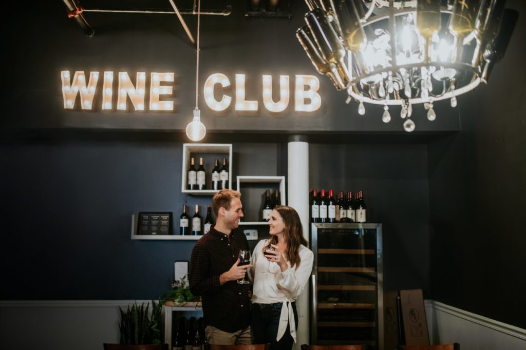 man and woman stand under wine club sign for their Louies Wine Dive engagement portraits