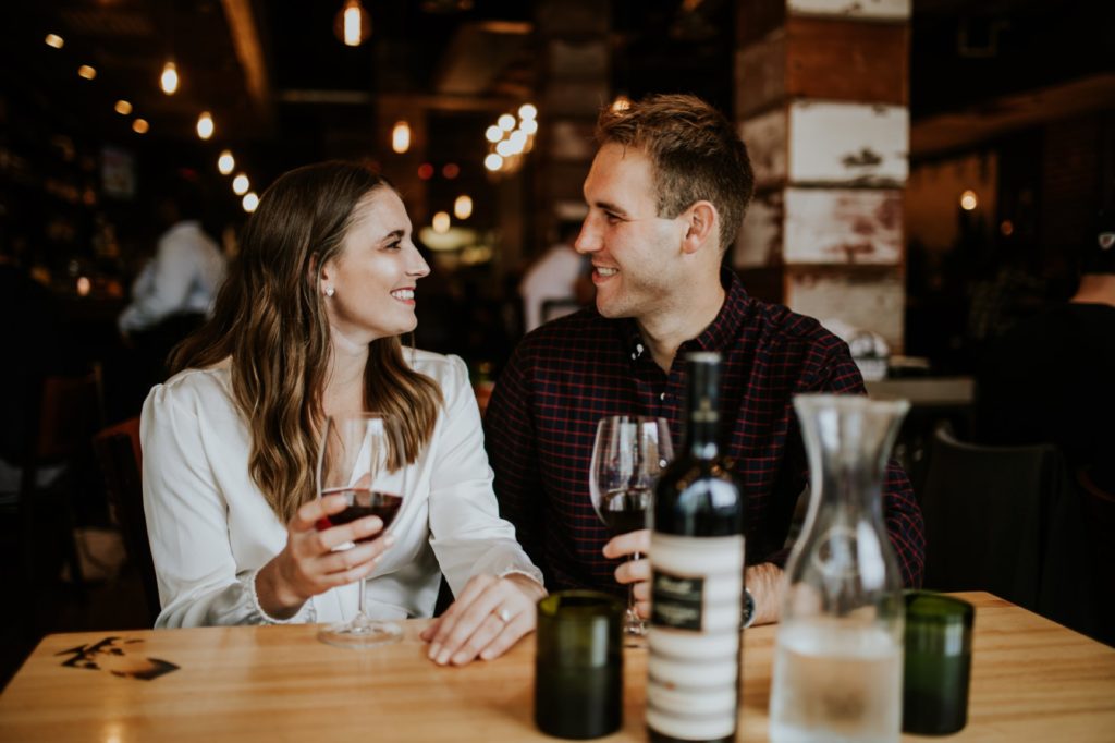 man and woman sit at table and drink wine together during their Louies Wine Dive engagement portraits