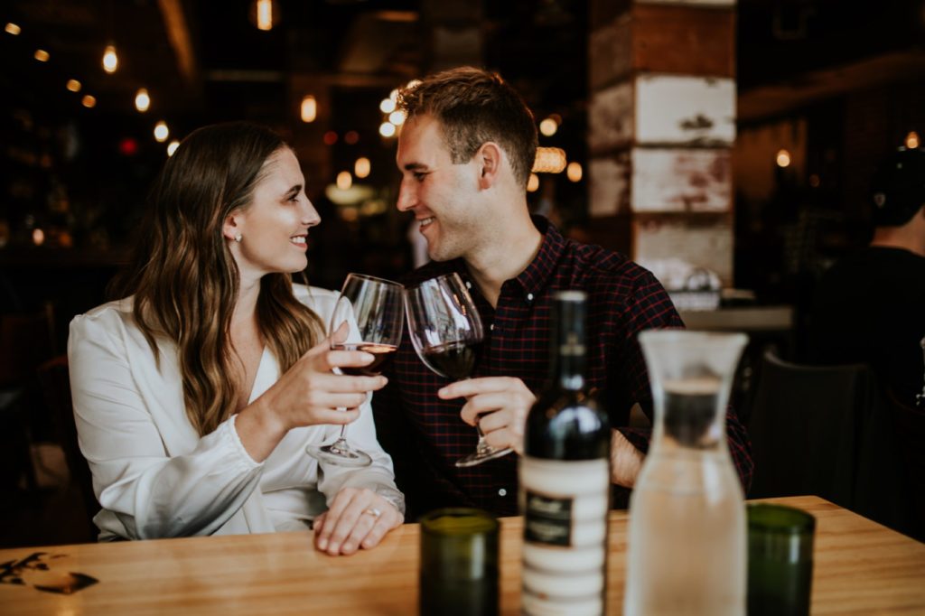man and woman clink wine glasses inside Louies Wine Dive for their engagement portraits