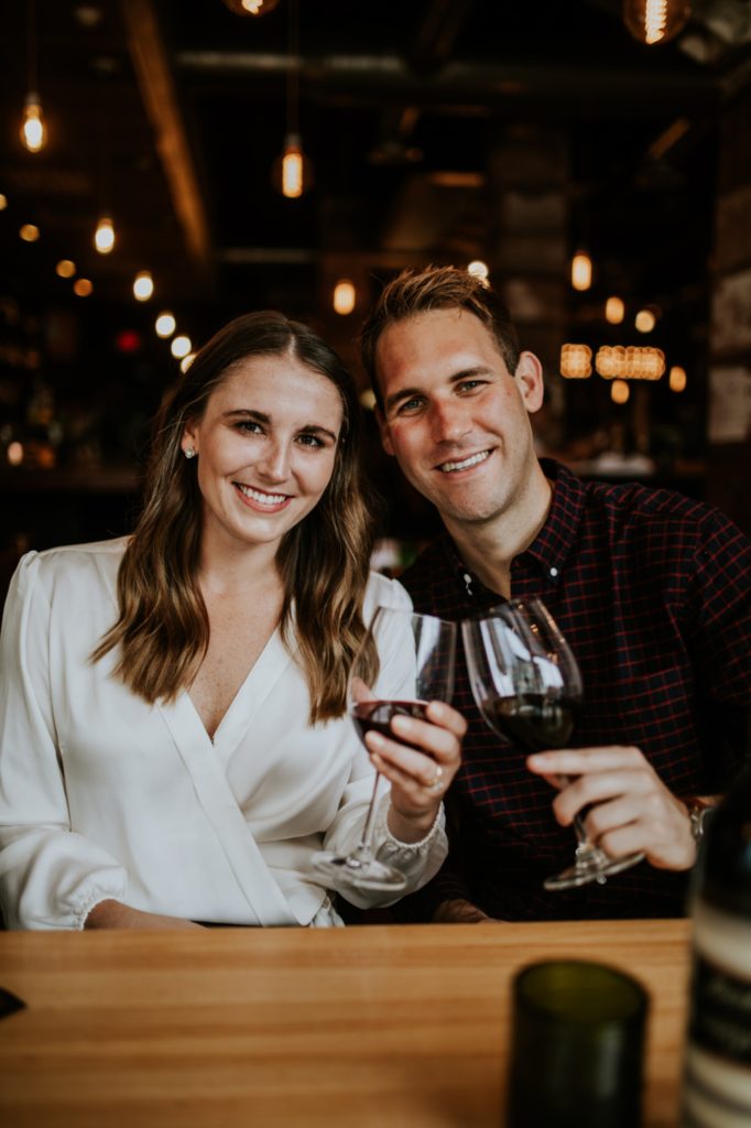 man and woman smile and look at camera with wine glasses in hand during their Louies Wine Dive engagement portraits