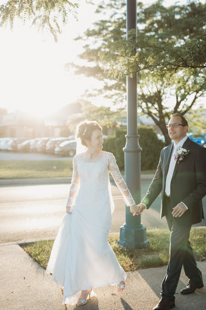 bride and groom hold hands and walk on sidewalk during golden hour at a carmel wedding