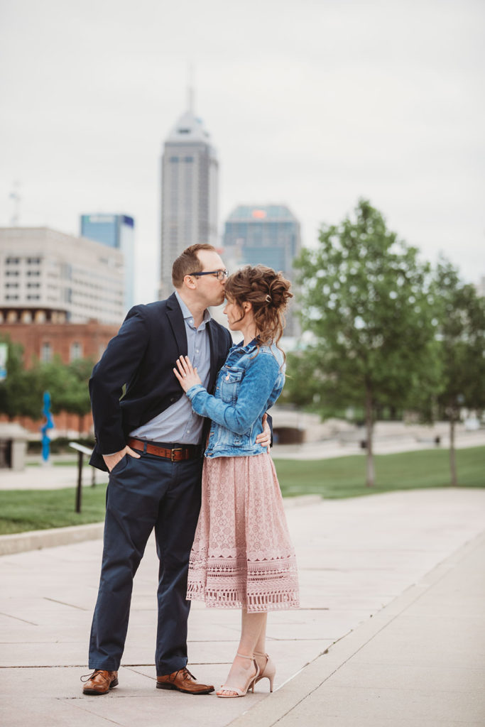 man kisses woman on forehead in front of indianapolis skyline during white river park engagement session