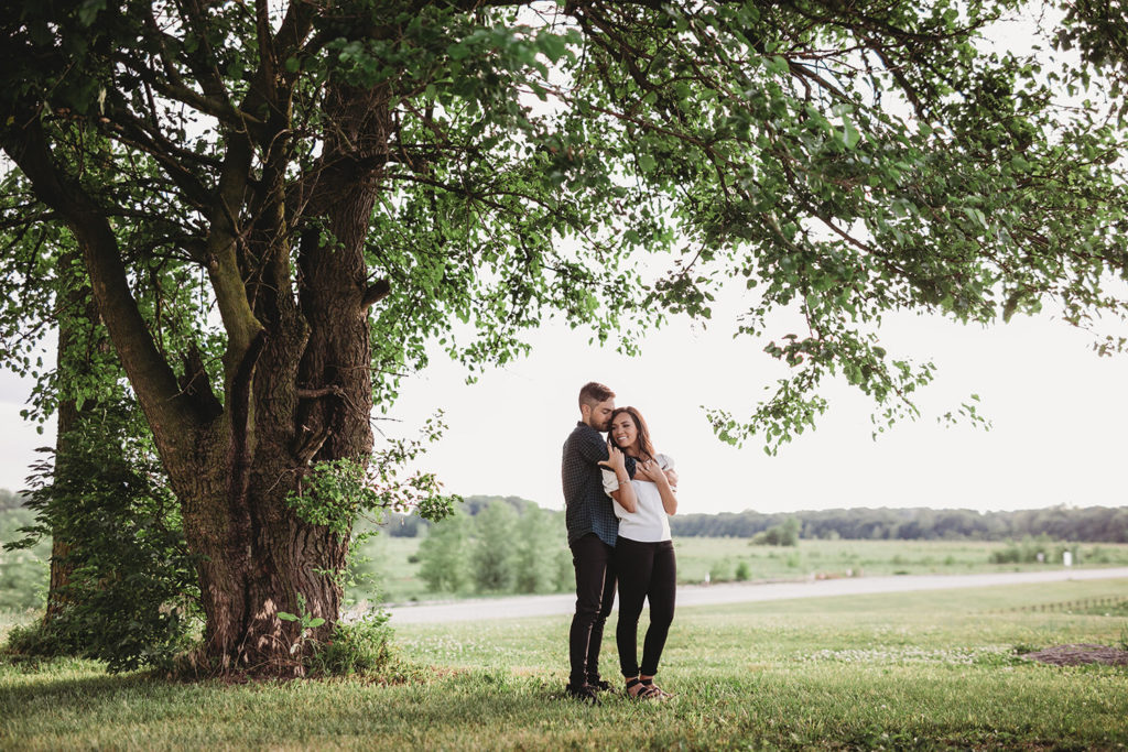 man hugs woman from behind under tree during their hamilton county engagement session