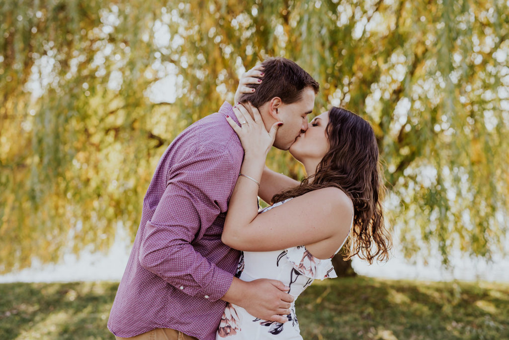 man and woman kiss passionately in front of weeping willow tree during their coxhall gardens engagement session