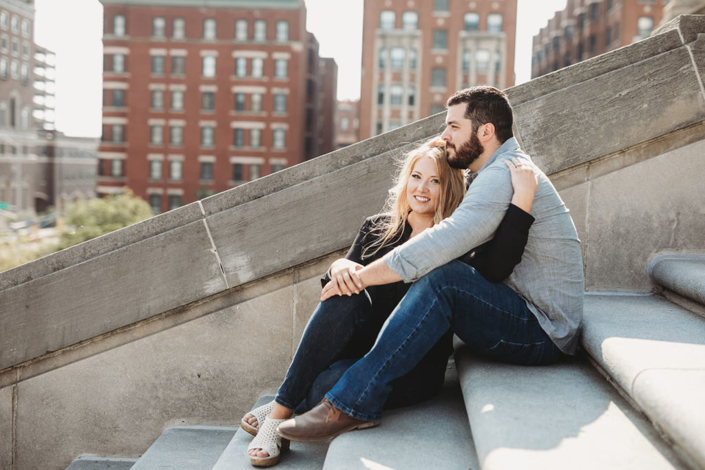 blonde woman and bearded man sit on steps of indiana war memorial during their downtown indianapolis engagement photos