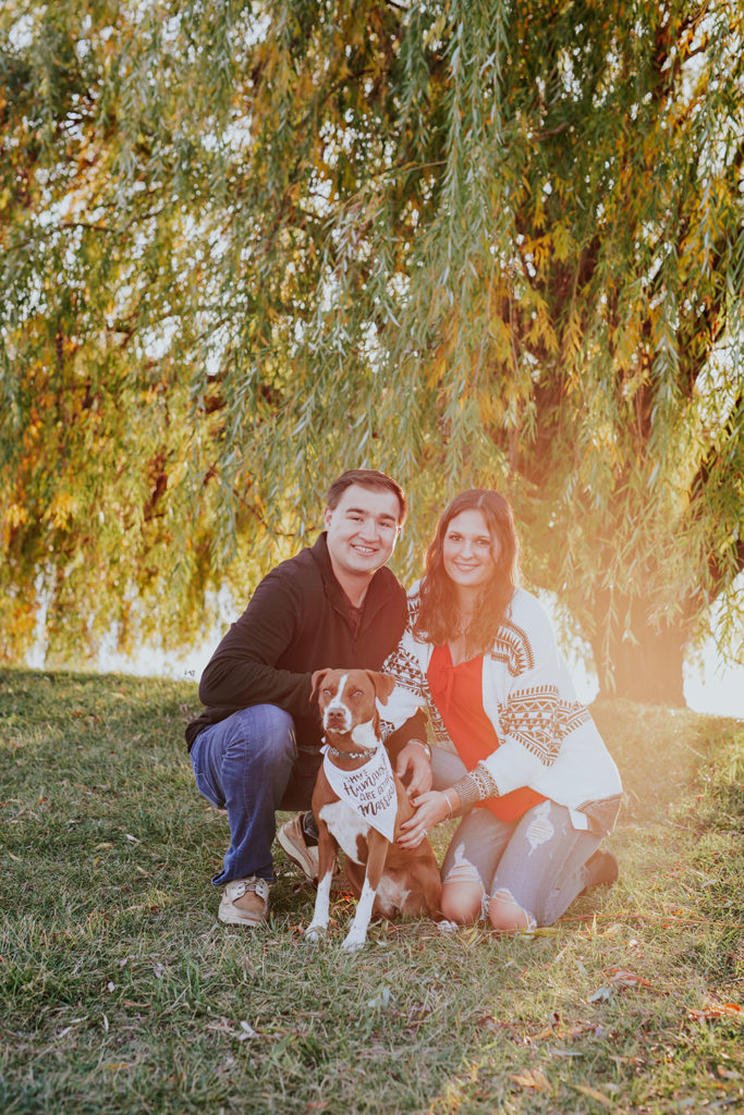 man and woman with dog at sunset in front of weeping willow tree at their coxhall gardens engagement session