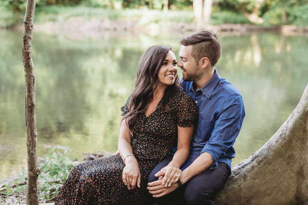 man and woman sit on exposed tree root in front of river during their hamilton county engagement