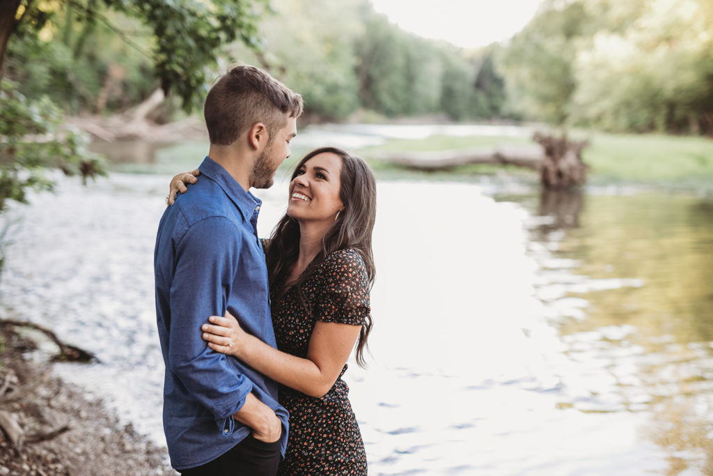 man and woman hug and smile in front of river at their Strawtown Koteewi Park engagement session