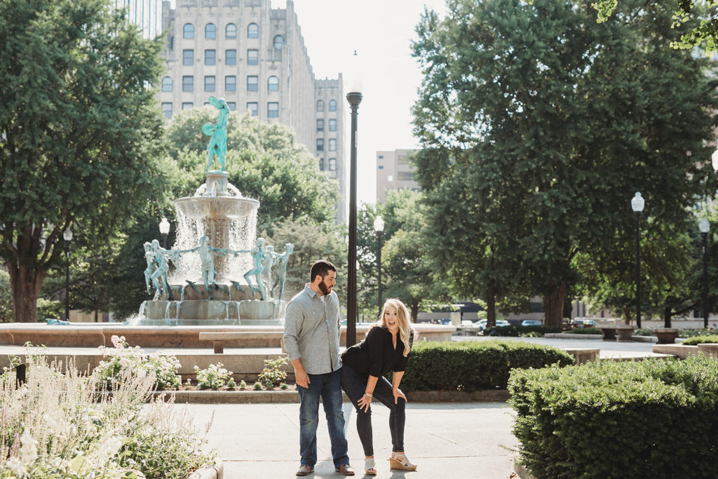 blonde woman pretends to sit on bearded man while he stands in park during their downtown indianapolis engagement photos