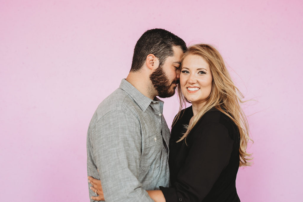 bearded man gets ready to kiss blonde woman in black on cheek in front of pink wall during their downtown indianapolis engagement photos