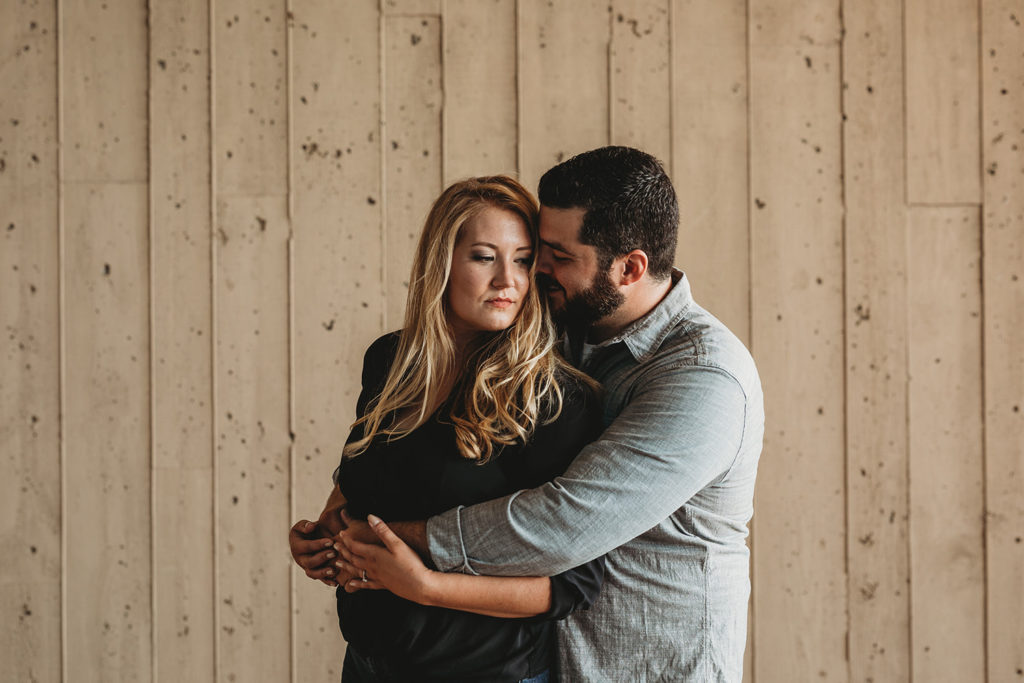 bearded man hugs blonde woman from behind in front of wood wall during their downtown indianapolis engagement photos