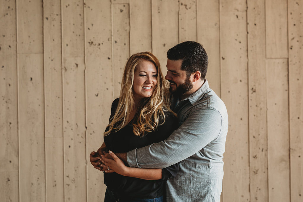 bearded man and blonde woman smile and laugh in front of wood wall