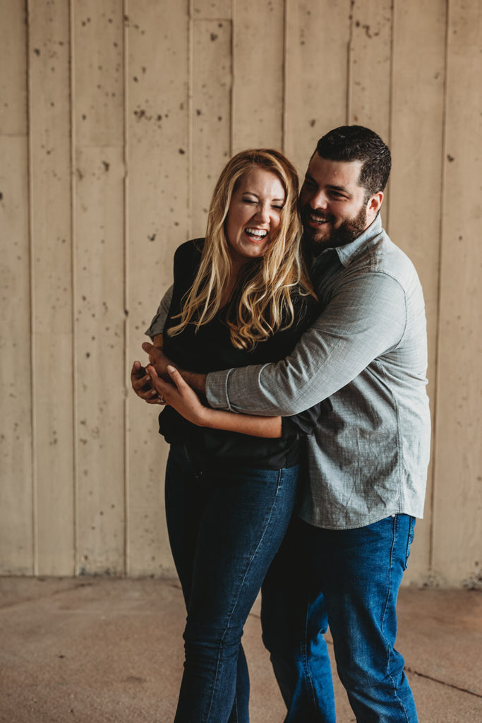 bearded man and blonde woman laugh uproarisly in front of wooden wall during their downtown indianapolis engagement photos