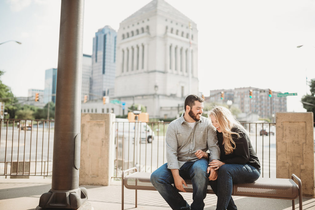 blonde woman in black and bearded man sit on bench in front of indiana war memorial in downtown indy laughing