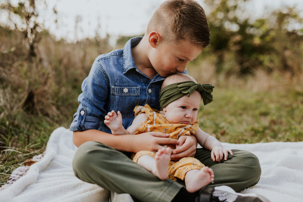 boy in denim shirt kisses baby sister in green headband on white blanket in Indy family photos