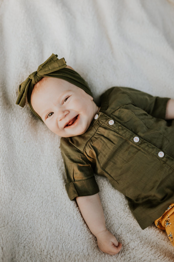 baby girl dressed in green shirt smiles on white blanket in Indy family photos
