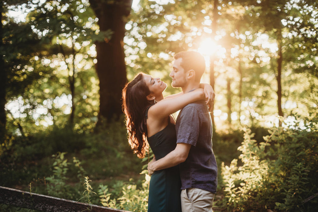 young couple hugging in woods in front of setting sun in woods at their Eagle Creek Reservoir Engagement