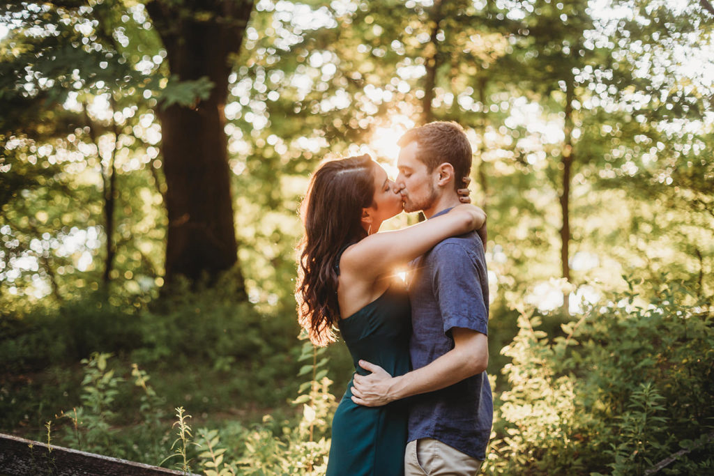 young couple kissing in front of sunset in woods at their Eagle Creek Reservoir Engagement